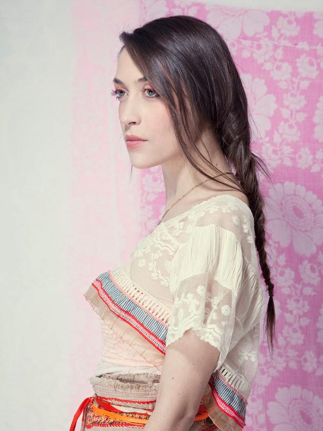 Modèle de profil devant un fond rose à motifs, corsage de dentelle écrue et ceinture tissée, teint mat et regard souligné pour la photographie de mode