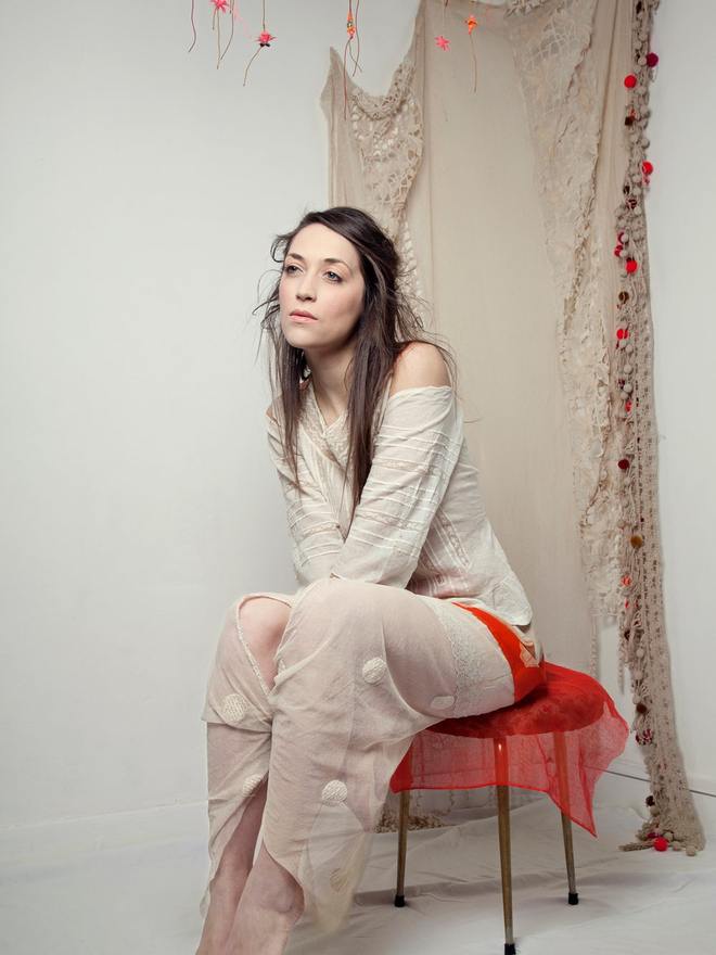 Modèle assise sur un tabouret dans un décor de studio aux voilages écrus et tulle rouge, maquillage de mode en tons neutres