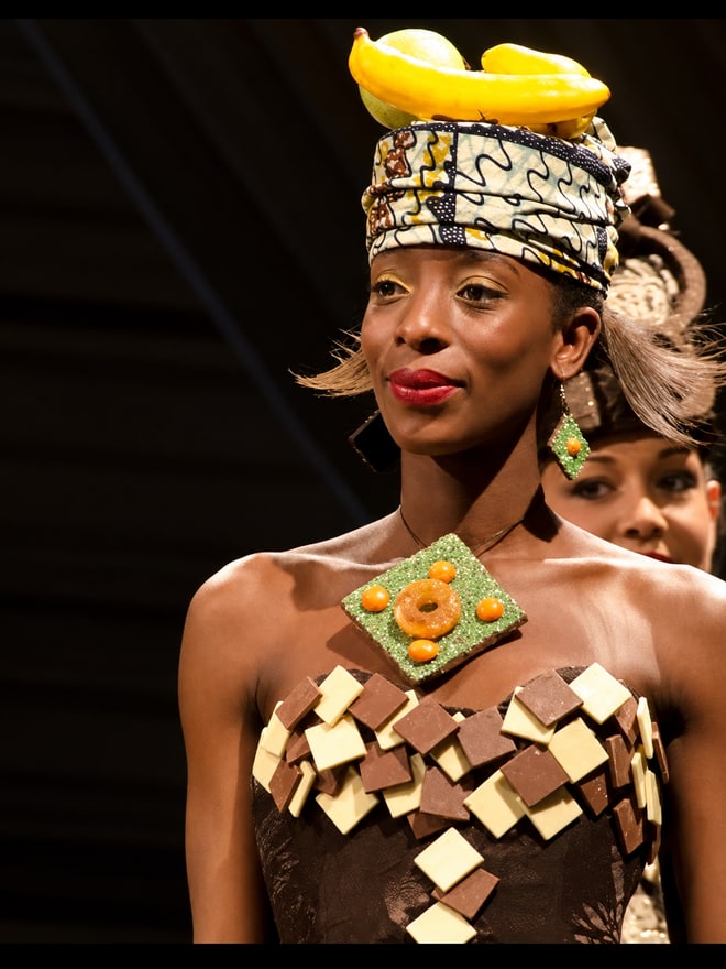 Mannequin en costume de défilé et coiffe jaune, maquillage lisible depuis la salle, présentation en lumière artificielle